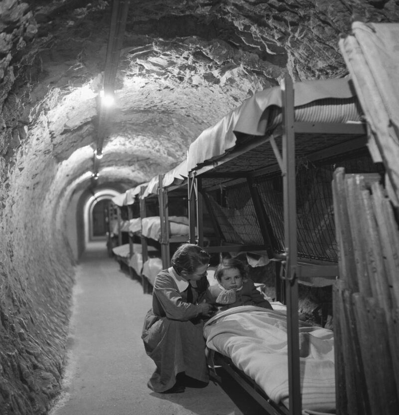 A black and white photograph of an old air raid shelter with a nurse caring for a child in a bunkbed 
